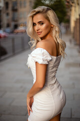 Beautiful girl in a white dress on the background of the city. A cute slender model with beautiful curls and a charming smile. Sunset light.