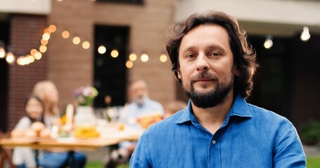 Portrait of Caucasian man with beard standing at yard smiling at camera. Male at family barbecue. Happy people sitting at dinner table on background.