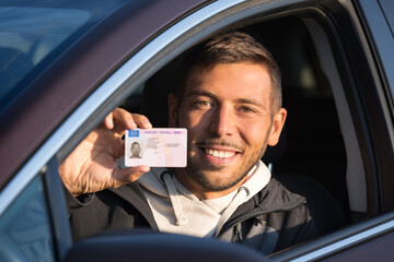 Attractive smiling young man proudly showing his driving license out of car window. Man has got driving license and feels very happy and excited. Ready to drive