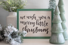 Closeup Of A Cute Christmas Sign And Miniature Trees Grouped On A White Table With Selective Focus