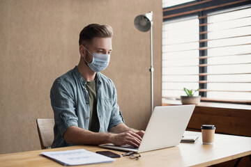 Young businessman working in office wearing medical protective face mask. Creative professional using laptop. Business, epidemic, pandemic, corona virus protection, healthy lifestyle, people concept