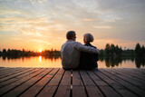 Fototapeta Morze - Romantic holiday. Senior loving couple sitting together on lake bank enjoying beautiful sunset.