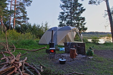 Beautiful camp site with metal wood-burning stove, camping tent, firewood and tall pine trees on a quiet summer evening in an empty forest clearing on river shore, outdoor tourism romantic lifestyle