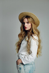 Wall Mural - Beautiful young long-haired woman in casual clothes and a straw hat posing in the studio on a gray background.