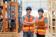 © amorn - Portrait of smiling Asian male and female warehouse worker in safety vest and helmet standing with arms crossed, holding clipboard in storage warehouse with shelf pallet spare parts parcel background
