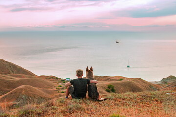 young man with his dog is sitting in the mountains and looking at the sea sunset. german shepherd dog travels with the owner. pets friendship and travel concept.