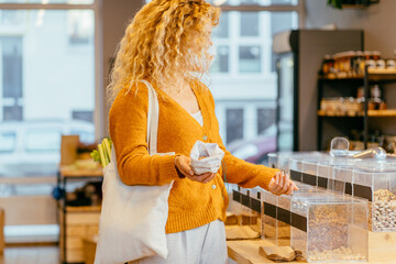 Wall Mural - Blond curly girl in yellow cardigan with cotton bag makes conscious shopping in zero waste shop. Woman buying food in plastic free grocery store. Sustainable small businesses. Minimalist lifestyle.