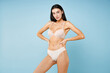 © ViDi Studio - Confident stunning beautiful attractive young brunette woman 20s in beige underwear showing sexy body standing posing with arms akimbo on waist isolated on pastel blue background, studio portrait.