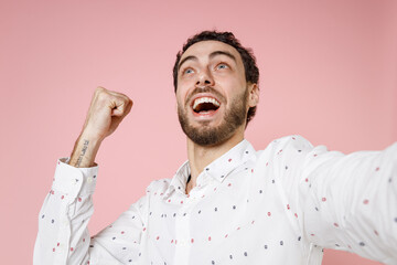 Wall Mural - Close up of excited young bearded man 20s in basic casual white shirt standing doing selfie shot on mobile phone doing winner gesture looking up isolated on pastel pink background studio portrait.