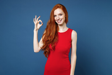 Wall Mural - Smiling pretty charming attractive young redhead woman 20s wearing bright red elegant evening dress standing showing OK gesture looking camera isolated on blue color wall background studio portrait.