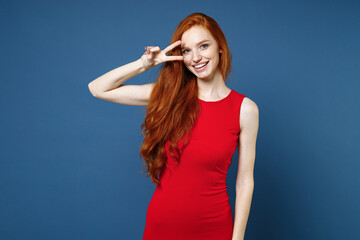 Wall Mural - Smiling pretty funny charming young redhead woman 20s wearing bright red elegant evening dress standing showing victory sign looking camera isolated on blue color wall background studio portrait.