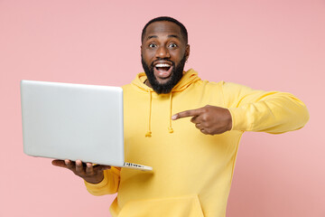 Excited funny young african american man 20s wearing casual yellow streetwear hoodie standing pointing index finger on laptop pc computer isolated on pastel pink color wall background studio portrait.