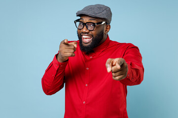 Canvas Print - Smiling cheerful young bearded african american man 20s wearing casual red shirt cap eyeglasses standing pointing index fingers on camera isolated on pastel blue color background studio portrait.