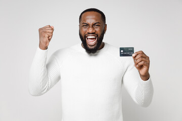 Happy joyful young african american man 20s wearing casual basic sweater standing doing winner gesture hold credit bank card looking camera isolated on white color wall background studio portrait.