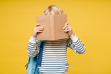 Llittle male kid teen boy 10s years old in striped sweatshirt backpack hold reading covering face with school book isolated on yellow color wall background, child studio portrait. Education concept.