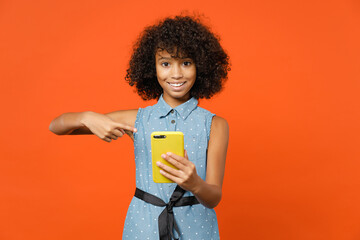 Smiling little african american kid girl 12-13 years old in basic denim dress pointing index finger on mobile cell phone typing sms message isolated on orange background . Childhood lifestyle concept.