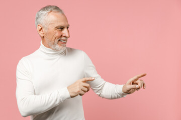 Smiling elderly gray-haired mustache bearded man wearing casual basic white turtleneck pointing index fingers aside on mock up copy space isolated on pastel pink color wall background studio portrait.