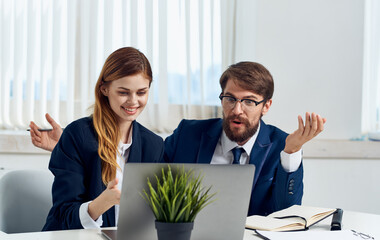 A woman and a man in suits look at a laptop monitor and flowers in a pot in the background