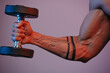 A close photo of a muscular arm which is doing bicep curls with a dumbbell under blue and red lights. A forearm of a man with a tattoo: Work it.