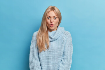 Wall Mural - Omg concept. Blonde scared woman with amazed expression keeps mouth opened being shocked by hearing unbelievable news wears knitted casual sweater isolated over blue background. Human reaction