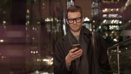 Fototapete - Man in eyeglasses with phone in hand on background of building with lights on. Handheld night shot of caucasian man texting in phone near business building with cars
