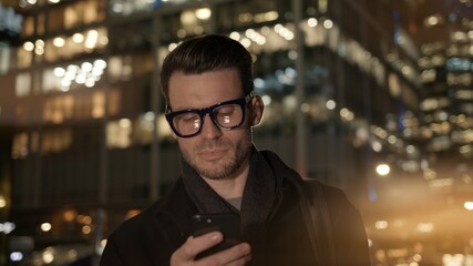 Fototapete - Man in eyeglasses with phone in hand on background of building with lights on. Gimbal night shot of caucasian man texting in phone near business building