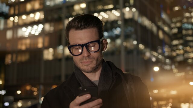 Man in eyeglasses with phone in hand on background of building with lights on. Gimbal night shot of caucasian man texting in phone near business building