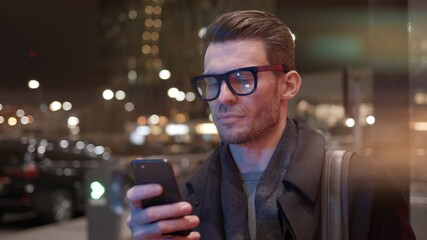 Fototapete - Man in eyeglasses with phone in hand on background of building with lights on. Gimbal night shot of caucasian man texting in phone near business building with cars