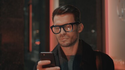 Fototapete - Man in eyeglasses with phone in hand on background of building with lights on. Handheld night shot of caucasian man texting in phone near business building with cars