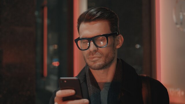 Man in eyeglasses with phone in hand on background of building with lights on. Handheld night shot of caucasian man texting in phone near business building with cars