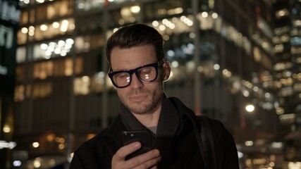 Fototapete - Man in eyeglasses with phone in hand on background of building with lights on. Gimbal night shot of caucasian man texting in phone near business building
