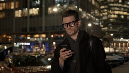 Fototapete - Man in eyeglasses with phone in hand on background of building with lights on. Gimbal night shot of caucasian man texting in phone near business building