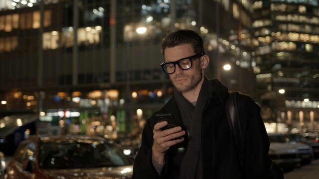 Man in eyeglasses with phone in hand on background of building with lights on. Gimbal night shot of caucasian man texting in phone near business building