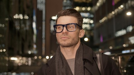 Man in eyeglasses standing on background of business skyscrapers, windows with lights. Portrait gimbal shot of businessman in coat standing near business centre downtown at night