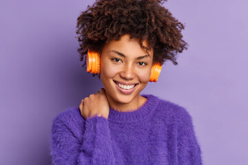 Poster - Headshot of good looking curly haired woman with Afro hair smiles gently touches neck looks positively at camera listens audio track via wireless stereo headphones isolated over purple background