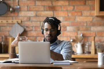 Serious young african ethnicity man in eyeglasses and modern wireless headset looking at computer screen, involved in video web camera call conversation, studying distantly or working remotely at home