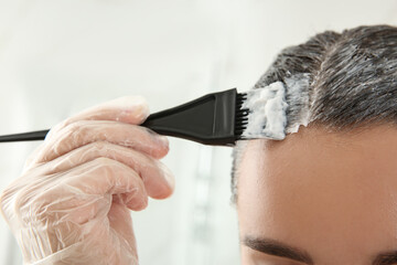 Wall Mural - Young woman applying dye on hairs against light background, closeup