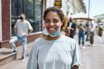 Wall Mural - Portrait of cheerful young african american woman with mask on her chin smiling at camera. People collecting their orders from the pickup point during coronavirus lockdown in the background