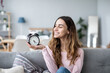 © BestForYou - Young smiling woman holds in hand alarm clock on the couch In a living room
