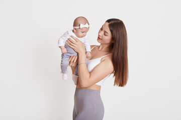 Wall Mural - Cheerful beautiful young woman in bra and grey leggins, holding baby girl in her hands and looking at her, brown haired female posing with child against white wall.