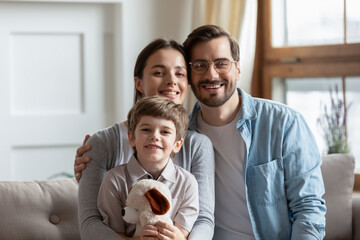 Wall Mural - Portrait of happy young family with little son hugging plush teddy bear in own living room. Smiling parents mom and dad with small boy child relax on couch at home, show unity and bonding.