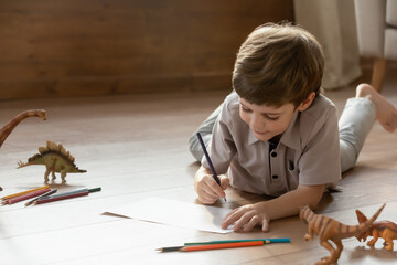 Cute little Caucasian preschooler boy lying on warm floor in living room drawing painting in album. Small child engaged in activity, playing having fun on weekend at home. Entertainment concept.