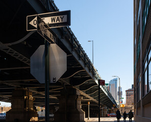 Two 'One Way' street signs intersecting under a bridge in dramatic light with silhouettes of pedestrians in the background