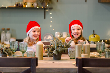 Two Happy cheerful Little girls in santa hat sitting at the festive table. Christmas time. New year 2021. Sisters having fun