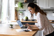 © fizkes - Serious young woman wearing glasses calculating finances, household expenses, confident businesswoman working with project statistics, using laptop and calculator, standing in kitchen at home