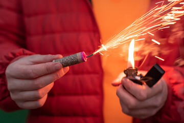 Wall Mural - Man in Red Jacked Lighting Up Firecracker in his Hand Using Gasoline Lighter. Guy Getting Ready for New Year Fun with Fireworks or Pyrotechnic Products