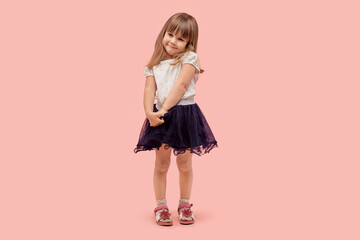 A cute, demure little girl in a black skirt and white tank top looks at the camera. Pink background.