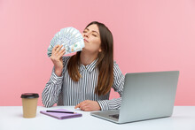 Excited Rich Business Woman Enjoying Smell Of Hundred Dollar Bills Holding Cash In Hand Sitting At Workplace, Crazy About Big Profit. Indoor Studio Shot Isolated On Pink Background