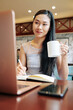 © DragonImages - Pretty young Chinese woman drinking mug of coffee when working on laptop in local cafe after classes in college