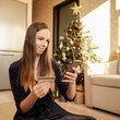 © Татьяна Максимова - Beautiful smiling brunette woman with mobile phone and credit card. Holidays sales, shopping online. Christmas tree on background, sunny day.